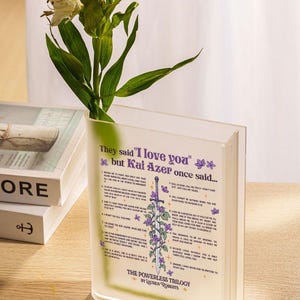 Op de afbeelding: Een heldere acrylvaas in de vorm van een boek met een citaat uit het boek "The Powerless Trilogy" van Lauren Roberts. Het citaat luidt "Ze zeiden "Ik hou van je" maar Kai Azer zei ooit..." en somt 12 redenen op waarom Kai Azer van iemand houdt. De vaas is bedrukt met een zwaard en bloemen.
