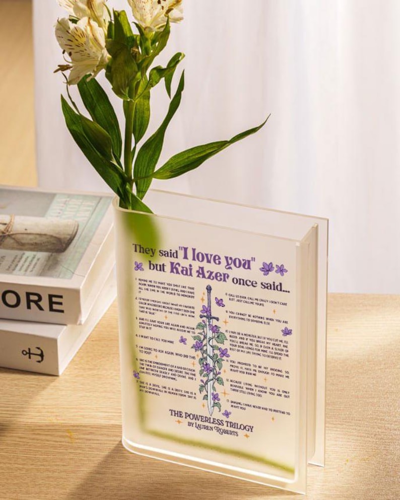 Op de afbeelding: Een heldere acrylvaas in de vorm van een boek met een citaat uit het boek "The Powerless Trilogy" van Lauren Roberts. Het citaat luidt "Ze zeiden "Ik hou van je" maar Kai Azer zei ooit..." en somt 12 redenen op waarom Kai Azer van iemand houdt. De vaas is bedrukt met een zwaard en bloemen.