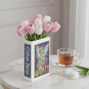 May include: A white ceramic vase shaped like a book, featuring a stained-glass design of a fairy and the text "Custom Name." The vase holds pink and white tulips. A cup of tea and two white tulips are on the table.