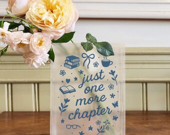 Puede incluir: Un jarrón rectangular transparente con el texto azul "just one more chapter" e ilustraciones de libros, flores y una taza de té. El jarrón contiene un ramo de rosas rosas y amarillas.