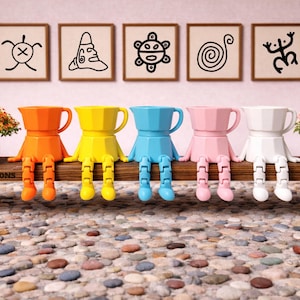 May include: A row of colorful coffee pot figurines with articulated legs, sitting on a wooden bench. The figurines are orange, yellow, blue, pink, and white. Above them are framed black and white abstract art pieces. Small bonsai trees flank the figurines.