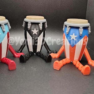 May include: Three 3D-printed conga drum figurines with legs. Each drum features a different color scheme: red, white, and blue; black and white; and orange, white, and blue. The drums have a star emblem and are sitting.