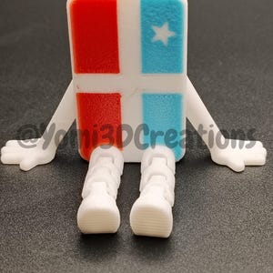 Puede incluir: Figura impresa en 3D con cuerpo rectangular, cuadrados rojos y azules, y una estrella blanca. Tiene brazos y piernas blancos, parecidos a un personaje de dibujos animados. La figura est&aacute; sobre una superficie oscura.