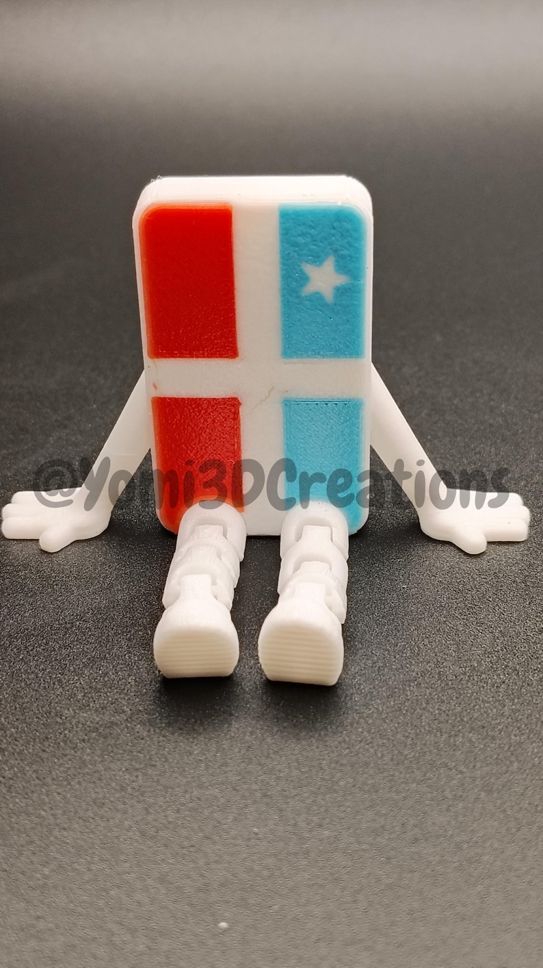Puede incluir: Figura impresa en 3D con cuerpo rectangular, cuadrados rojos y azules, y una estrella blanca. Tiene brazos y piernas blancos, parecidos a un personaje de dibujos animados. La figura est&aacute; sobre una superficie oscura.