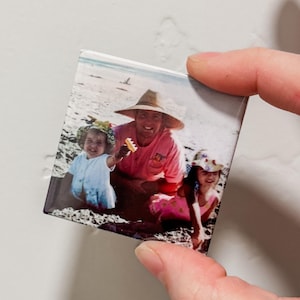 Op de afbeelding: Een kleurenfoto van een gezin van drie op een strand. De vader draagt een roze shirt en een strohoed. De twee dochters dragen hoeden en jurken. De oudste dochter houdt een klein speelgoed vast.