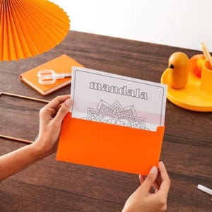 Può includere: Una busta arancione contiene una pagina da colorare con la parola "mandala" e un disegno floreale dettagliato. Una lampada e un blocco note arancioni abbinati sono su una superficie di legno scuro, insieme a un vassoio giallo con matite e un uccello decorativo.