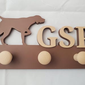 May include: Brown wooden coat rack with three hooks and a silhouette of a brown dog. The rack has the letters "GSP" in tan lettering.