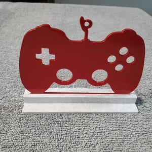 Puede incluir: Silueta roja de un mando de videojuegos, montada sobre una base texturizada blanca y gris. El mando tiene una cruceta, botones y un lazo en la parte superior. Un artículo decorativo para jugadores.
