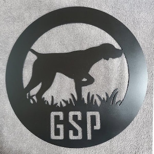 Puede incluir: Silueta negra de metal de un Braco Alemán de Pelo Corto en posición de caza, con las letras GSP debajo del perro. La silueta está sobre un fondo circular negro.