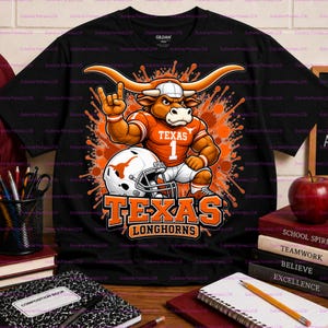 Könnte beinhalten: Ein schwarzes Kurzarm-T-Shirt mit einer Grafik eines Cartoon-Longhorn-Stiers, der ein orangefarbenes Trikot mit dem Text TEXAS 1 trägt und einen Football-Helm hält, vor einem orangefarbenen Farbspritzer-Hintergrund mit dem Text TEXAS LONGHORNS.