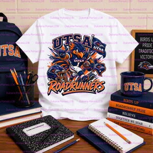 Op de afbeelding: Een wit t-shirt met een afbeelding van een blauwe en oranje vogelmascotte in sportkleding die een football vasthoudt, met de tekst UTSA ROADRUNNERS. Het ontwerp bevat een footballhelm en een vlag in levendige blauwe en oranje kleuren.