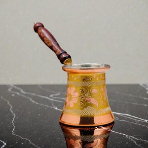 Könnte beinhalten: Eine türkische Kaffeekanne aus Kupfer und Gold mit einem Holzgriff. Der Topf hat rosa und goldene Blumen- und Blättermuster. Der Griff ist dunkelbraun mit einem abgerundeten Ende.