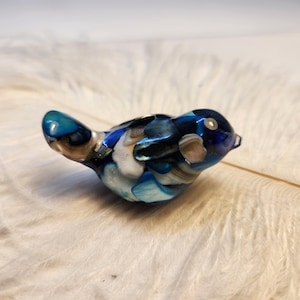 Puede incluir: Pequeña figura decorativa de pájaro con cuerpo redondeado y pico puntiagudo. El pájaro es principalmente azul, con detalles blancos y marrones. La figura descansa sobre una superficie blanca y suave, posiblemente plumas.