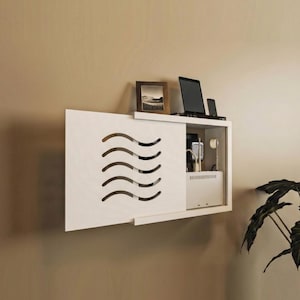 May include: A white, wall-mounted cabinet with a sliding door featuring a wave-like cutout design. The cabinet holds a laptop, phone, picture frame, and other electronics. The cabinet is mounted on a beige wall.