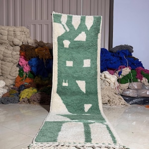 Op de afbeelding: Een rechthoekig, handgeweven tapijt met een groen en wit geometrisch ontwerp. Het tapijt heeft een centraal groen patroon met witte accenten en een franje langs de onderrand. Het tapijt wordt verticaal weergegeven.