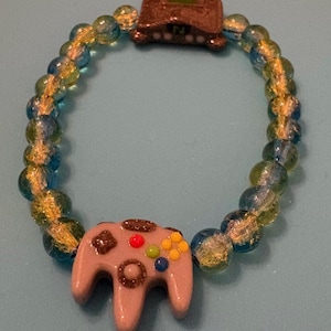 May include: A bracelet featuring a light gray controller charm with colorful buttons and a brown console charm. The bracelet is strung with blue and yellow glass beads, creating a playful accessory.