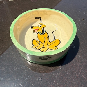 May include: A round ceramic pet bowl with a cream-colored interior featuring a hand-painted yellow cartoon dog with black ears and a green collar. The exterior rim is painted green, and a single black paw print is visible on the side of the bowl.