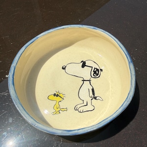 Peut inclure: Un bol rond en céramique avec un intérieur couleur crème présentant une illustration peinte à la main de Snoopy et Woodstock portant des lunettes de soleil. Le bol a une finition mouchetée et un bord peint en bleu, avec une surface lisse et vernie.