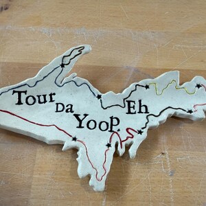 May include: A ceramic map of the Upper Peninsula of Michigan, with the words "Tour da Yoop Eh" in black lettering. The map features red, black, and yellow lines, and black star accents. The map is off-white and sits on a wooden surface.