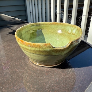 May include: A handmade ceramic bowl with a wide, shallow design. The bowl features a glossy green interior and a textured, earthy brown rim. The exterior has a mottled green and brown glaze, creating a natural, organic aesthetic. The bowl is approximately 12 inches in diameter.