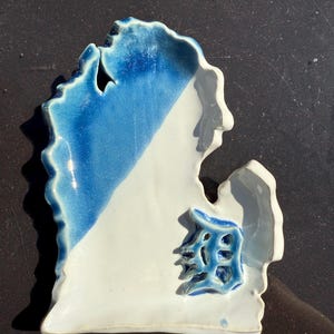 Könnte beinhalten: Eine Keramikschale in Form des US-Bundesstaates Michigan, mit einer diagonalen Teilung aus blau-weißer Glasur. Auf der weißen Seite ist ein blaues Emblem zu sehen. Die Schale ist handgefertigt und hat eine glänzende Oberfläche.