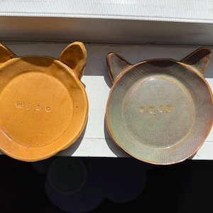 May include: Two ceramic cat-shaped dishes, one yellow with the word "miso" inscribed and the other green with the word "onigiri" inscribed.