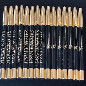 May include: A close-up of twelve black eyeliner pencils with gold caps. The pencils are labelled "Salon Atash" in gold lettering.