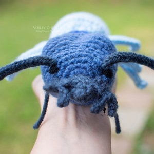May include: A crocheted blue and white bug with black antennae. The bug is being held by a hand.