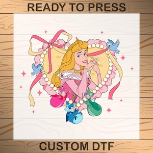 May include: A pink heart-shaped design with a princess in a pink dress and a crown. The princess is surrounded by three fairies and two birds. The design includes a pearl necklace and a yellow ribbon. The text "READY TO PRESS" is at the top and "CUSTOM DTF" is at the bottom.