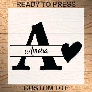 May include: Black graphic design featuring the letter "A" with the name "Amelia" in cursive script, a heart, and the text "READY TO PRESS" and "CUSTOM DTF". The design is on a white background with a wood grain border.