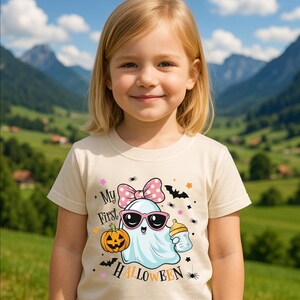 May include: A beige t-shirt featuring a cartoon ghost wearing sunglasses and holding a bottle. The shirt has the text "My First Halloween" and a pink bow with white polka dots. A jack-o'-lantern and bats are also featured.