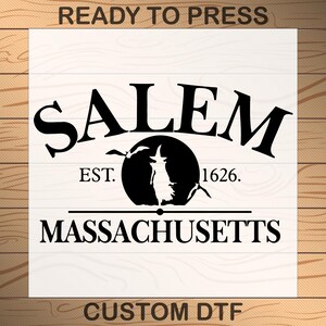 May include: A white square transfer sheet featuring black text and graphics. The text reads READY TO PRESS at the top, SALEM, EST. 1626., MASSACHUSETTS, and CUSTOM DTF at the bottom. A central black circle depicts a witch silhouette and two birds.