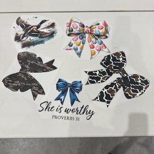 May include: A collection of decorative stickers featuring various designs. Included are a flying duck, several bows with colorful patterns, and a bow with a camouflage design. The text "She is worthy" is also present.