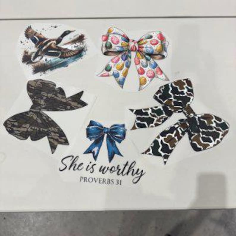 May include: A collection of decorative stickers featuring various designs. Included are a flying duck, several bows with colorful patterns, and a bow with a camouflage design. The text "She is worthy" is also present.