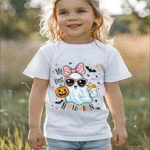 May include: White t-shirt featuring a cartoon ghost wearing sunglasses and a pink bow, holding a pumpkin and a baby bottle. The text "My First Halloween" is printed on the shirt. The shirt is designed for children.