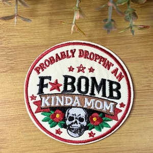 May include: Circular embroidered patch with the text "PROBABLY DROPPIN' AN F BOMB KINDA MOM". The design includes a skull, red stars, and red flowers with green leaves. The border is red.