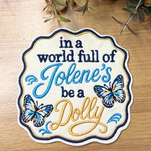 May include: An embroidered patch with a scalloped edge featuring the text in a world full of Jolene's be a Dolly. The design includes two butterflies with blue, white, and yellow wings, with dark blue and gold thread accents on a cream background.