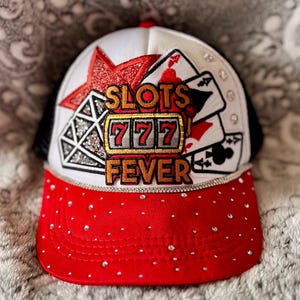 May include: A white and red trucker hat with a black mesh back. The front of the hat features a red star, a diamond, playing cards, and a slot machine with the words "Slots Fever" and "777". The hat is decorated with rhinestones.