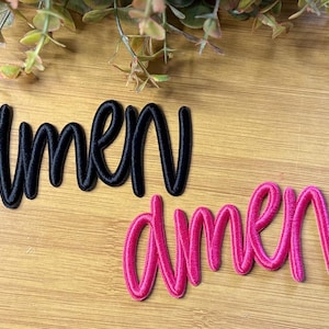 May include: Two embroidered patches featuring the word amen in a cursive script font. One patch is black and the other is bright pink. Both patches have a raised, stitched texture and are designed to be applied to fabric surfaces.