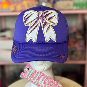 May include: A purple trucker hat with a large bow design. The bow is white with gold glitter accents and purple outlines. The hat has a mesh back and a curved brim. There are small glitter accents on the brim.