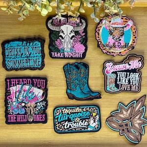 May include: A collection of embroidered patches with a Western theme. Designs include a steer skull, cowboy boots, and phrases like "Take No Shit" and "Rowdy & Reckless." The patches feature vibrant colors and detailed stitching.