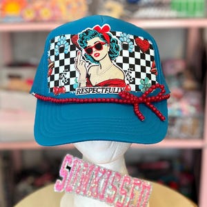 May include: A turquoise trucker hat with a mesh back and a red beaded accent. The front features a graphic of a woman with sunglasses and the word "RESPECTFULLY" below. The hat is displayed on a mannequin head.