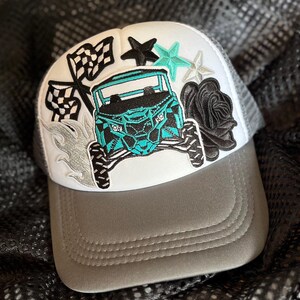 May include: A gray and white trucker hat with a teal and black off-road vehicle, black and white checkered flags, a black and white star, a silver star, and a black rose.