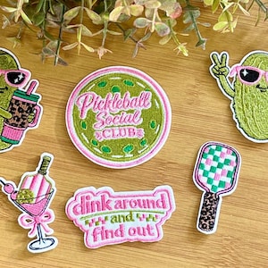 May include: A collection of embroidered patches with a pickleball theme. Includes patches of pickles wearing sunglasses, a cocktail, a paddle, and text that says "Pickleball Social Club" and "dink around and find out".