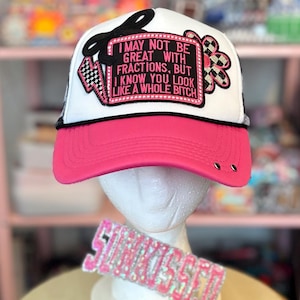 May include: A white and pink trucker hat with the text "I MAY NOT BE GREAT WITH FRACTIONS, BUT I KNOW YOU LOOK LIKE A WHOLE BITCH." The hat has a pink brim and a black mesh back. A decorative headband and a gold hand-shaped heart are also visible.