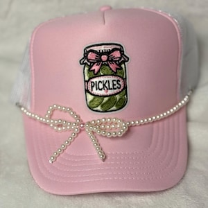 May include: Pink trucker hat with a white mesh back and a pearl beaded bow. The front of the hat features an embroidered patch of a pickle jar with the word "PICKLES".