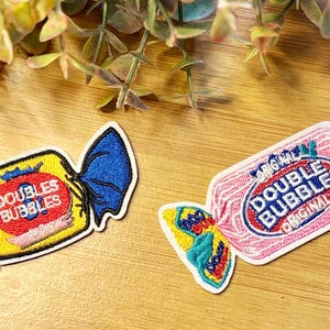 May include: Two embroidered patches shaped like wrapped candies. One is yellow with blue accents and reads "Doubles Bubbles." The other is pink with blue and yellow accents and reads "Double Bubble Original."