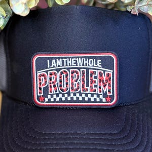 May include: Black trucker hat with a mesh back and a patch that reads "I AM THE WHOLE PROBLEM" in white and red leopard print lettering. The patch has a red border and a checkered pattern at the bottom.