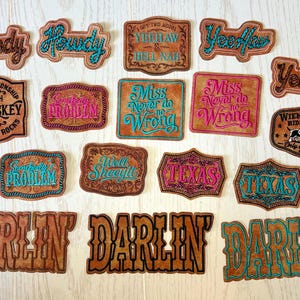 May include: A collection of brown leather patches with various embroidered phrases and designs. Phrases include "Howdy," "Darlin'," and "Texas." Some patches have turquoise or pink accents, and others feature a dog or whiskey theme.