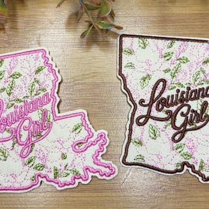 May include: Two embroidered patches shaped like the state of Louisiana, featuring a white background with a pink and green floral pattern. One patch has a pink border and pink text reading Louisiana Girl, while the other has a brown border and brown text.